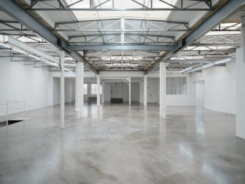 Empty, bright hall with a concrete floor, white walls, columns, and an exposed steel beam ceiling.