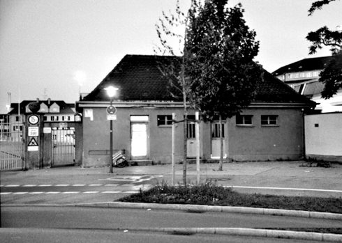 Schwarzweißaufnahme von Das KloHäuschen, der Eingang und ein Fenster sind hell erleuchtet, der Platz davor ist leer.