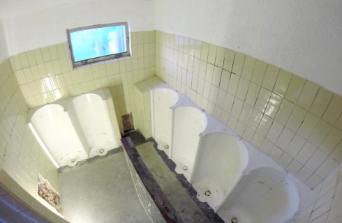 Innenraum von Das KloHäuschen mit gefliesten Wänden und mehreren Urinalen, ein kleines Fenster befindet sich an der oberen Wand.