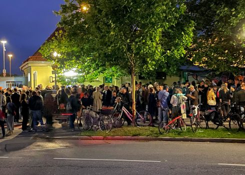 Große Menschenmenge vor Das KloHäuschen am Abend, das Gebäude ist von Bäumen und Straßenlaternen umgeben, viele Fahrräder stehen davor.