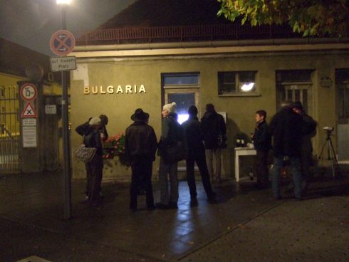 Menschen versammeln sich abends vor Das KloHäuschen, an der Fassade steht der Schriftzug „Bulgaria“, ein Fenster ist von innen beleuchtet