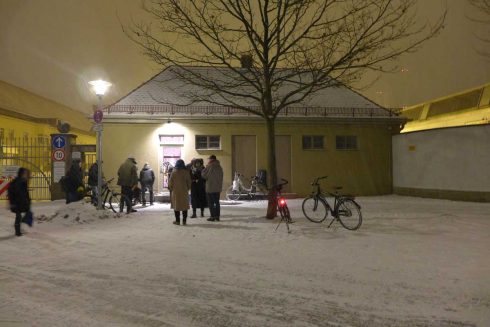 Außenansicht von Das KloHäuschen bei Nacht im Winter, mehrere Personen stehen vor dem Gebäude und blicken auf ein hell erleuchtetes Fenster.
