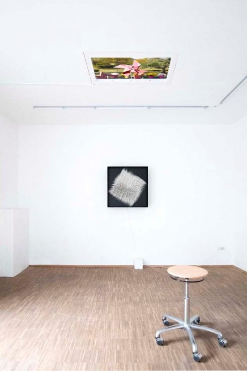 Heller Ausstellungsraum mit Kunstwerken an Wand und Decke sowie einem rollbaren Hocker.