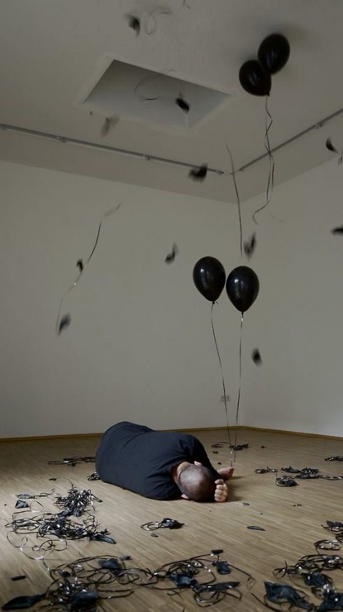 Person liegt zusammengerollt auf dem Boden eines Ausstellungsraums, umgeben von schwarzen Luftballons und herabfallenden Bänder