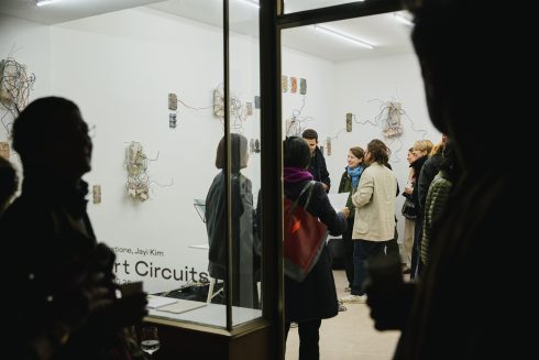 Blick durch eine Glasfront in einen Ausstellungsraum mit Wandarbeiten und Besucher*innen.
