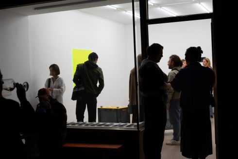 Blick durch eine Glasfront in einen Ausstellungsraum mit Wandarbeiten und Besucher*innen.