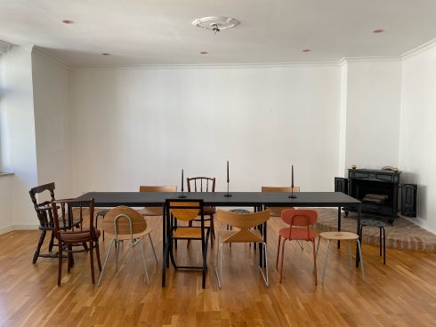Raum mit langem schwarzen Tisch, unterschiedlich gestalteten Stühlen, Kerzenhaltern auf dem Tisch und einem Kamin an der rechten Wand.