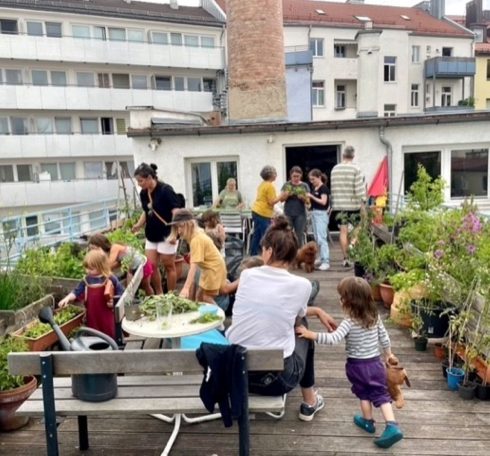 Mehrere Personen halten sich auf einer Dachterrasse zwischen Pflanzkübeln, Tischen und Bänken auf.