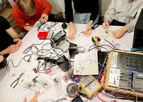 Mehrere Personen arbeiten an einem Tisch mit Kabeln, elektronischen Bauteilen, Werkzeugen.