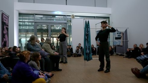 Zwei Personen führen Bewegungen in einem Ausstellungsraum aus, das Publikum sitzt im Halbkreis um die offene Fläche.