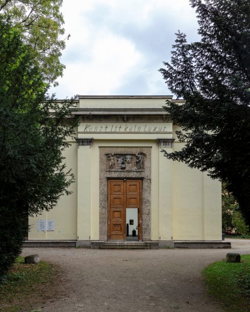 Frontansicht des Kunstpavillons mit klassischer Fassade, geöffnetem Holztor und Bäumen links und rechts