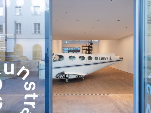 Blick durch eine Glasfront in einen Ausstellungsraum mit einem länglichen Flugzeugrumpf auf Rollen, beschriftet mit dem Wort „LIBERTÉ“.