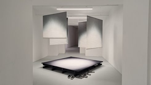 Minimalistischer Ausstellungsraum mit Podest, hängenden Lamellen und abgelegten Schuhen.