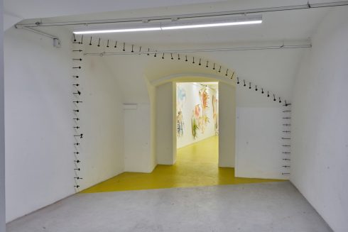 Ausstellungsraum mit weißer Architektur, gelbem Bodenbereich und Wandinstallation aus Nägeln.
