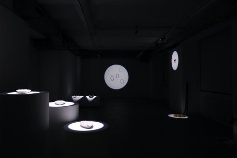 Dunkler Ausstellungsraum mit beleuchteten Podesten und kreisförmigen Projektionen an den Wänden.