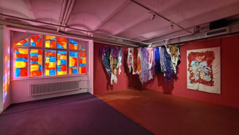 Ausstellungsraum mit farbigen Stoffarbeiten, Wandbild und bunt verglastem Fenster.