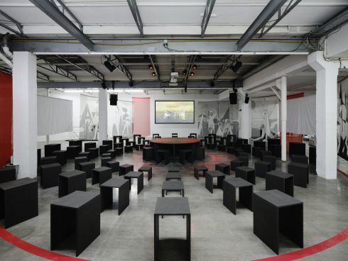 Exhibition space with many black stools, a round table in the center, and a projection on the wall.
