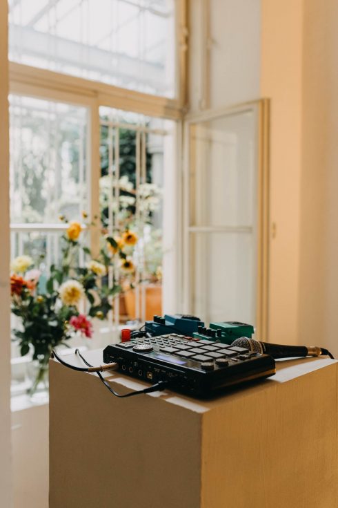 Audio-Setup mit Sampler, Effektgeräten und Mikrofon auf einem Sockel vor einem geöffneten Fenster, dahinter Blumenarrangements
