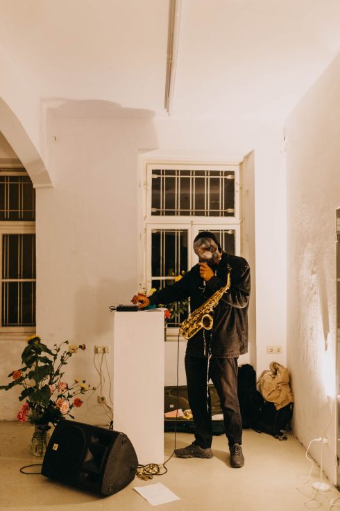 Eine Person steht neben einem Sockel und hält ein Saxophon, während sie ein kleines Audiogerät bedient; auf dem Boden liegen Kabel, ein Lautsprecher und ein Blatt Papier, im Hintergrund ein vergittertes Fenster mit Blumen.