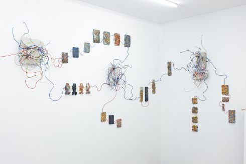 Ausstellungsansicht: Wandinstallation aus farbigen, reliefartigen Modulen, verbunden durch frei geführte Drähte.
