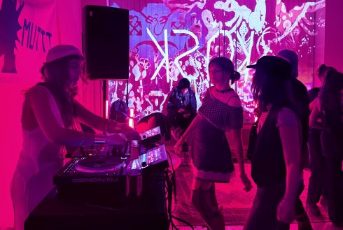 DJ-Performance in pinkem Licht, Besucherinnen tanzen vor einer projizierten Wandgrafik.