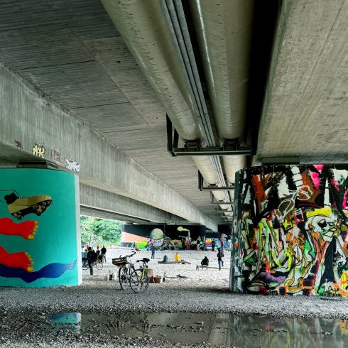 Blick unter eine Brücke mit bemalten Betonpfeilern, Fahrrädern, Personen und auf dem Boden verteilten Materialien.