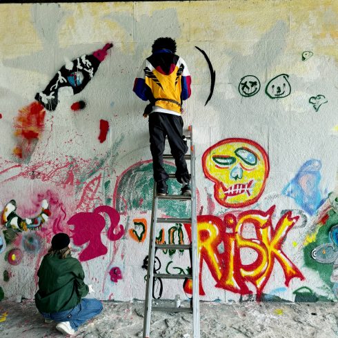 Zwei Personen arbeiten an einer Wand mit Graffiti, eine steht auf einer Leiter, eine kniet davor auf dem Boden.