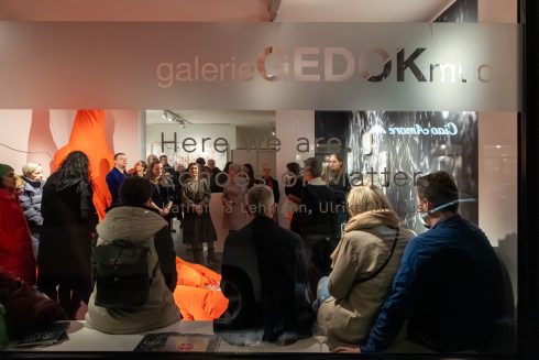 Blick durch eine Glasfront in den Ausstellungsraum der galerie GEDOK München während einer Vernissage: Zahlreiche Besucher*innen stehen und sitzen dicht gedrängt vor einer Installation mit großformatigen orangefarbenen Textilien; an den Wänden sind weitere künstlerische Arbeiten zu sehen, Schriftzüge auf der Scheibe überlagern die Szene.