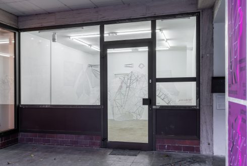 Blick durch eine Glasfront in einen weißen Ausstellungsraum mit einer großformatigen, netzartigen Skulptur aus Draht oder Stäben.