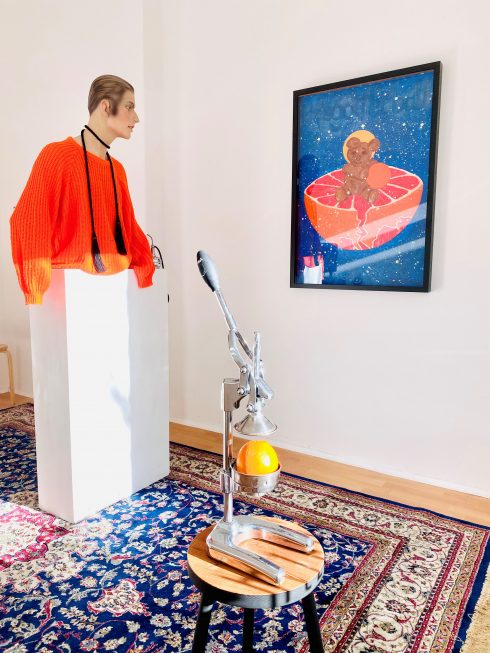 Ausstellungsansicht mit Büste im orangefarbenen Pullover neben einer Zitruspresse mit Orange und gerahmtem Bild.