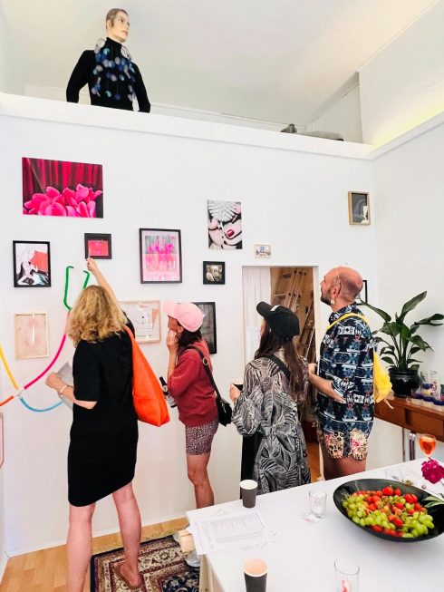 Ausstellungsbesuch mit mehreren Personen vor einer Wand mit kleinen Kunstwerken. Über der Wand steht eine Figur.
