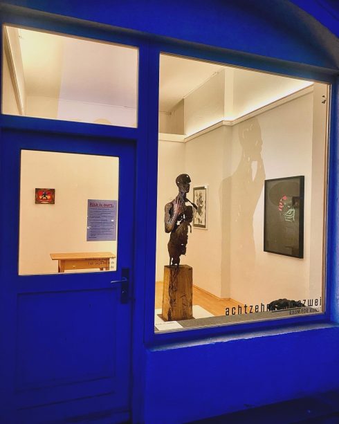 Blick von außen in einen beleuchteten Ausstellungsraum mit Skulptur auf Sockel und gerahmten Werken an den Wänden.