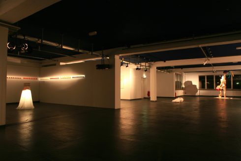 Großer, abgedunkelter Ausstellungsraum mit mehreren installativen Arbeiten und gezielter Beleuchtung; einzelne Skulpturen sind räumlich verteilt.