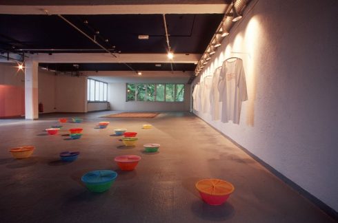 Großer Ausstellungsraum mit farbigen Schalen, die rasterartig auf dem Boden verteilt sind, sowie textilen Arbeiten an der Wand; keine Personen im Raum.