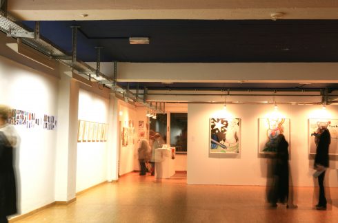 Innenraum einer Ausstellung mit gerahmten Arbeiten an den Wänden; mehrere Besucher*innen bewegen sich durch den Raum, teils als Bewegungsunschärfen sichtbar.