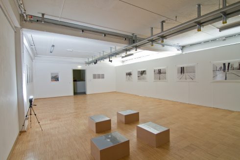 Heller Ausstellungsraum mit Holzboden, mehreren wandhängenden Fotografien und vier niedrigen Sockeln in der Raummitte; keine Personen anwesend.