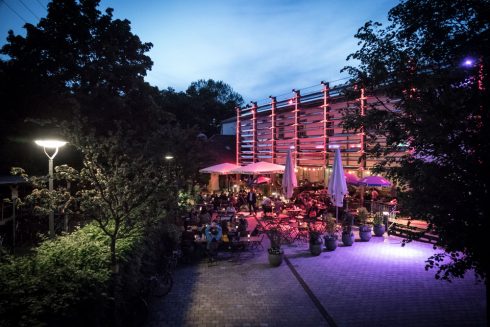 Abendliche Außenansicht beleuchtetem Gebäude und Sitzbereich im Freien, zahlreiche Besucher*innen sitzen und stehen zwischen Tischen unter Bäumen.