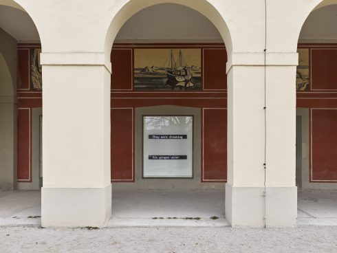 Arkadengang mit drei hellen Rundbögen; in der mittleren Nische ein Schaufenster mit leuchtender Textarbeit vor rot bemalter Wand und einem Wandbild mit Boot darüber.