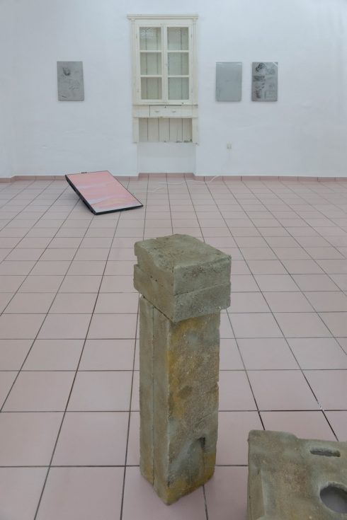 Innenansicht eines Ausstellungsraumes. Im Vordergrund steht eine eckige Steinskulptur. Im Hintergrund hängen Bilder und ein altes Holzfenster and er Wand.